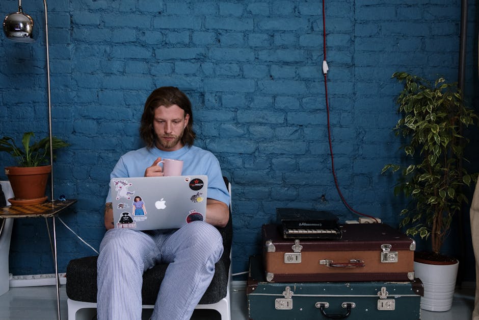 Bearded man with long hair sitting indoors, working on a sticker-covered laptop and holding a mug.