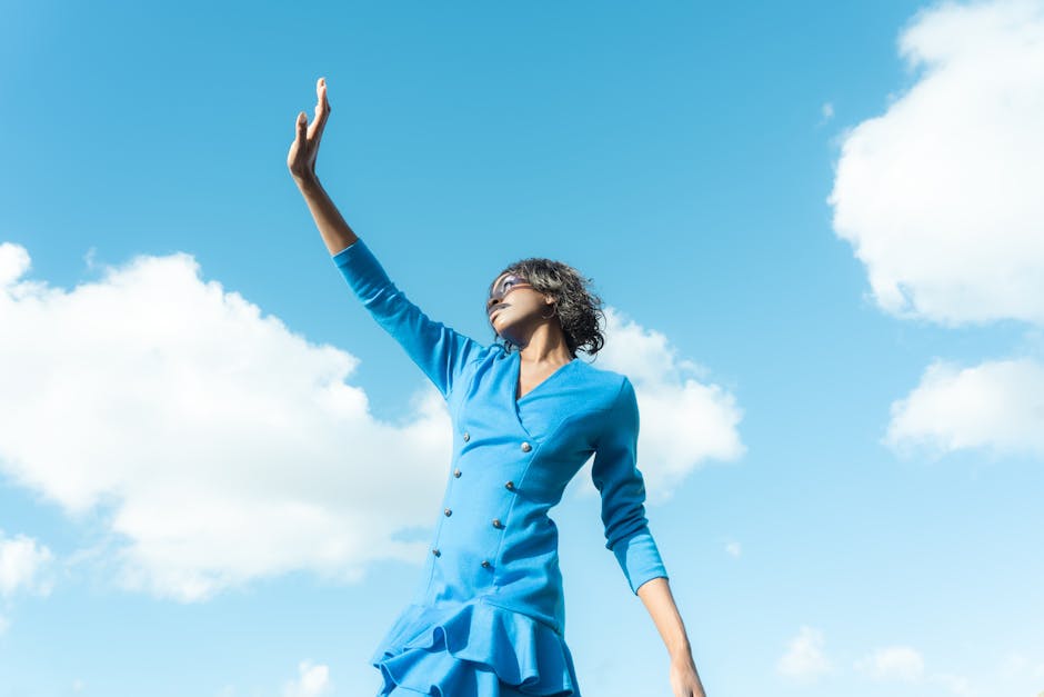 Stylish woman in blue dress reaching up, set against a bright blue sky.