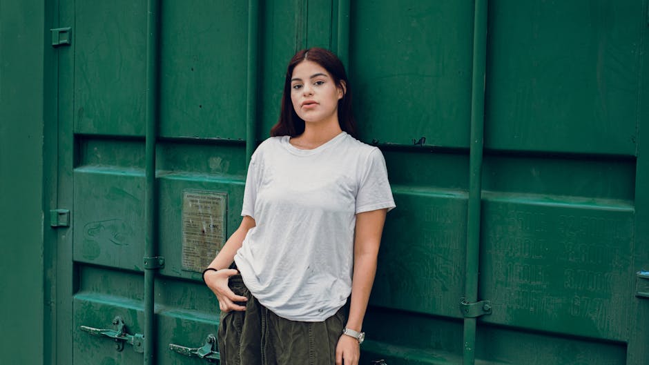 Young woman in casual attire standing against a green metal background outdoors.