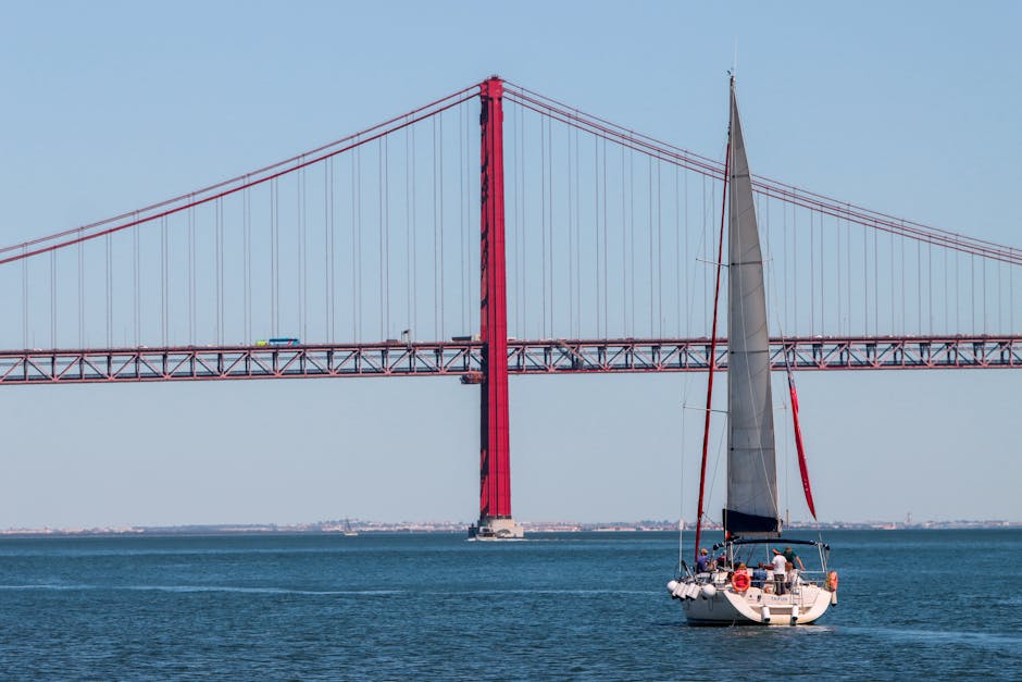 Sailboat under the iconic 25th of April Bridge in Lisbon, Portugal, on a clear day.
