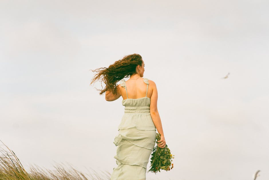 A woman with flowing hair stands calmly with a bouquet on a windy coastline, embodying tranquility and freedom.